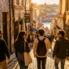 Quatro turistas descem uma rua estreita de calçada, ao pôr do sol, com o rio e a cidade ao fundo
