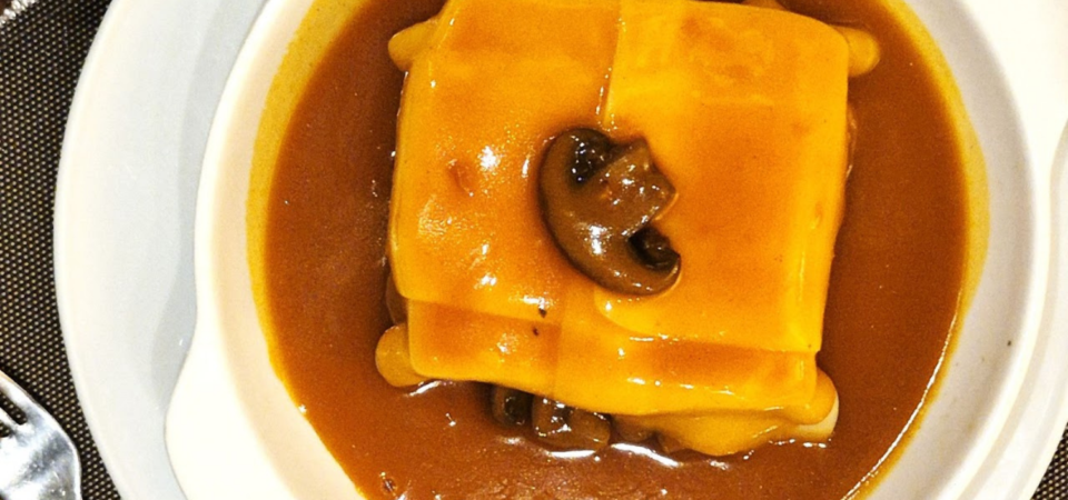 Prato de francesinha e cogumelos, coberto por uma fatia de queijo derretido